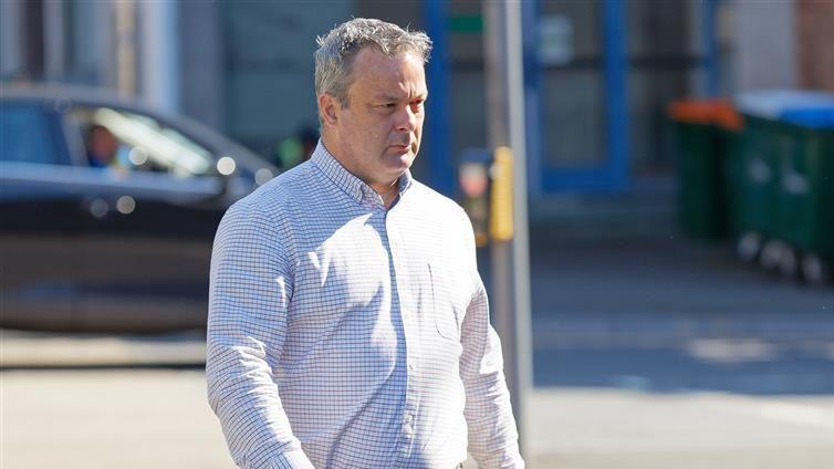 Christopher Elias walking in the sun to the court, he is wearing a white and grey checked shirt, has grey hair and has a furrowed brow.