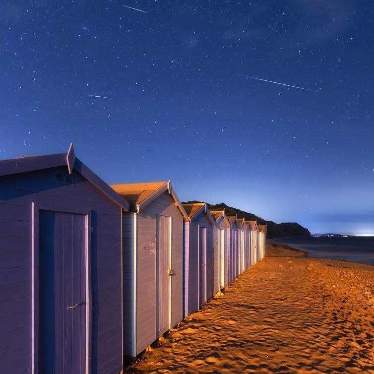 Meteors shimmering in the sky above Charmouth Beach, where purple and white beach huts can be seen in the foreground 