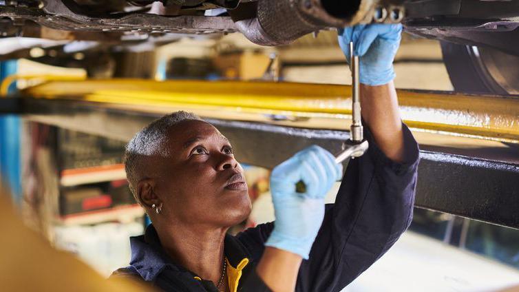 Young black woman stands underneath car, using a tool to fix the undercarriage. She has short grey hair, and is wearing blue overalls and light-blue surgical gloves. 