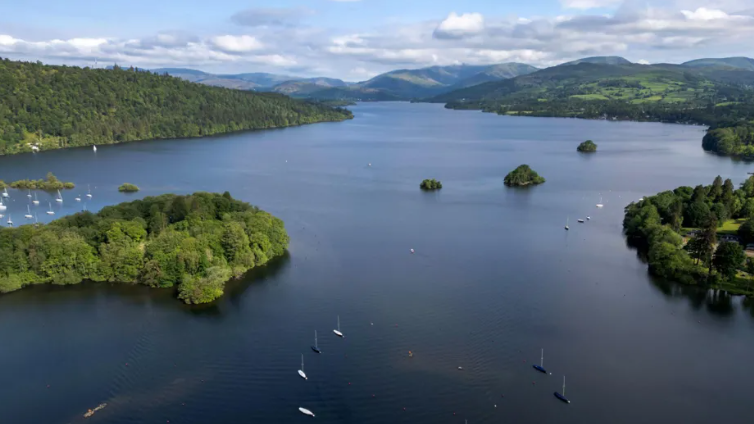 An aerial view of a large lake with numerous boats floating on it. It is surrounded by green hills and trees.