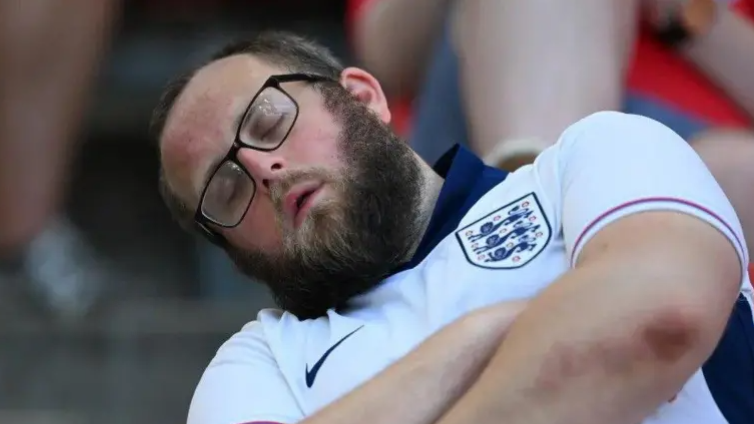 A man asleep. He is wearing glasses and a white England shirt. 