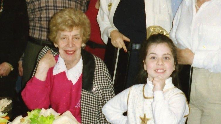 A young girl sits next to an elderly woman in a pink jumper. They are at a party and are sitting at a table of food, with other people stood behind them