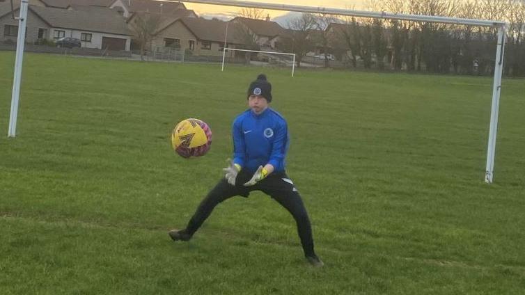 Adam Golebiewski training as a goalkeeper before his injury, wearing dark trousers, a blue top and a dark hat, preparing to catch a yellow football, among goalposts.