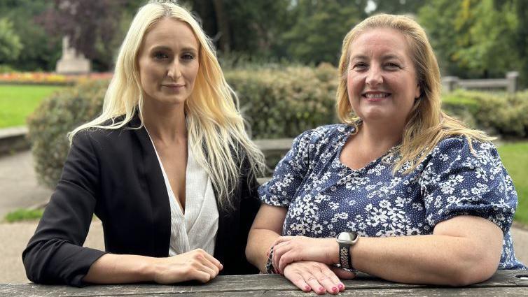 Two women seated at what appears to be a picnic table. The one on the left has long blonde hair and is wearing a black jacket and white blouse. The other has long fair hear and a blue and white short sleeved top