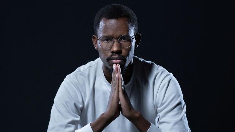 Nelson Amenya, in a white top, holds his hands in front of him in a prayer position as he stares into the camera.