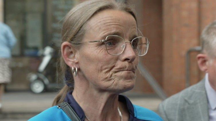 Joanne Moody standing outside a redbrick building, wearing large specs on her face, hoop earrings, a necklace, and a blue sports jersey. The background is softly blurred and includes steps leading to the building entrance and a partially visible mobility scooter.