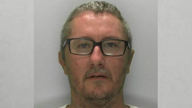 A mugshot of a man in his early 40s with short grey hair, short facial hair and black rimmed glasses, who is staring blankly