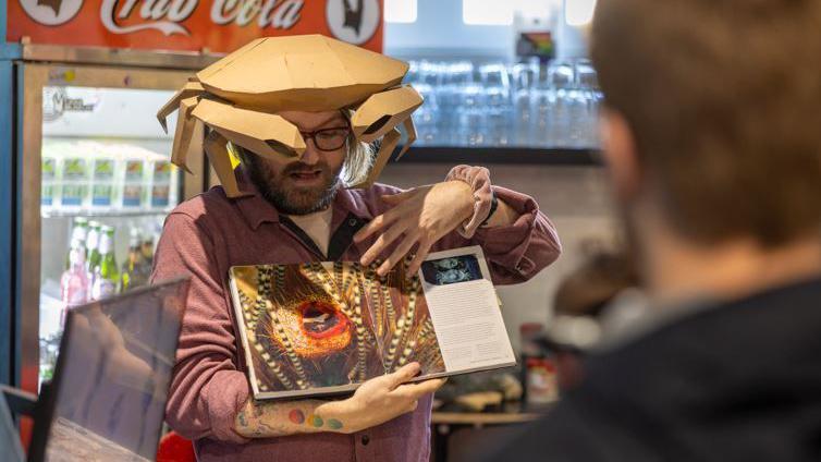 A member of staff at Crab Museum in Margate wearing a crab hat explaining details about crabs from a book