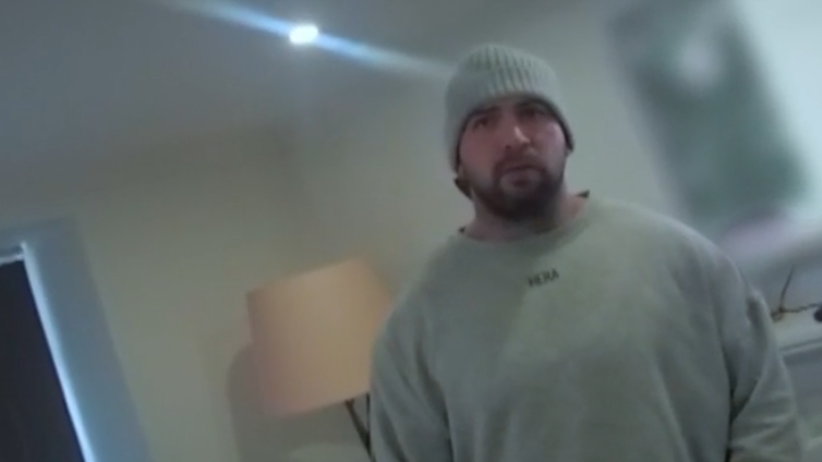 Bodycam footage of a man in a grey sweather and grey beanie hat stands in his living room with a lamp and a television behind him