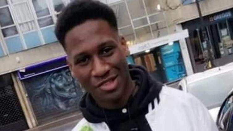 A young man with black afro hair. He is wearing a black hoodie with a white jacket. Behind him is a load of shops. 