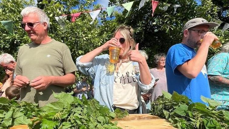 Bethan Hodges drinking cider and giving the thumbs up during the contest. She is standing in between two men and next to a table piled up with stinging nettles.