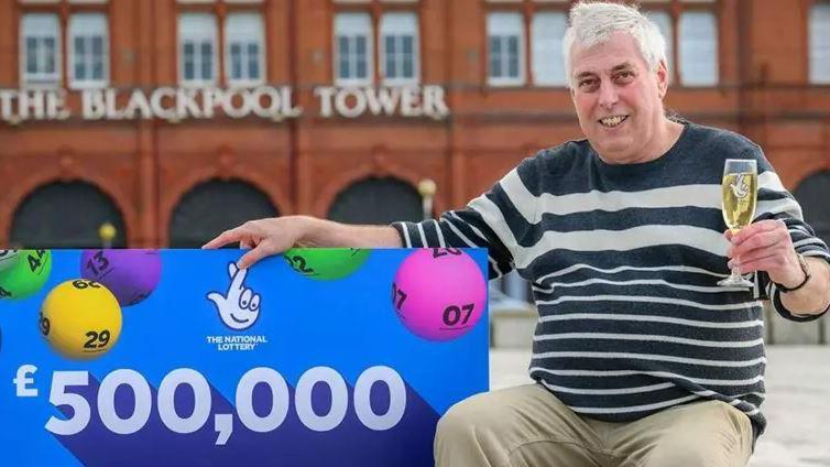 A man sits to the right of the camera, holding a glass of bubbly, while holding a huge blue cheque with the writing '£500,000' on. The Blackpool Tower building can be seen in the background 