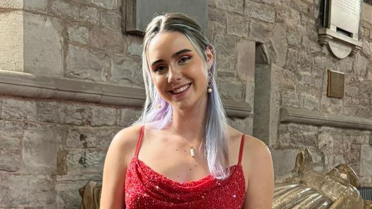 Megan Hastings smiles at the camera, she is wearing a red sparkly dress and has long brown hair which is dyed lilac. She is standing in a church. It is a head and shoulders shot of her. 