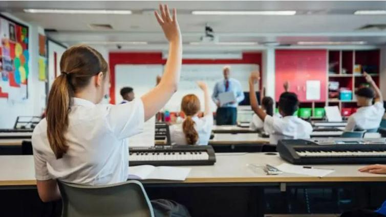 An image of a girl at the back of a classroom with her hand in the air. The image is taken from behind.