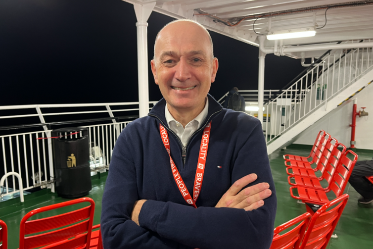 Man with bald head stands on ferry deck in front of red chair with a dark morning sky