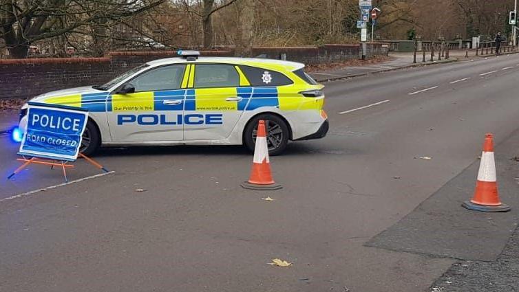 Street view of Riverside Road- with a white and yellow police car vertically placed on the road with a police sign in front of it and orange cones closing the road 