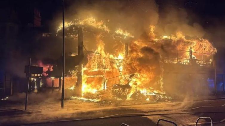 A huge fire engulfs a building.  It is night time and the sky is dark.  The building is next to a road.