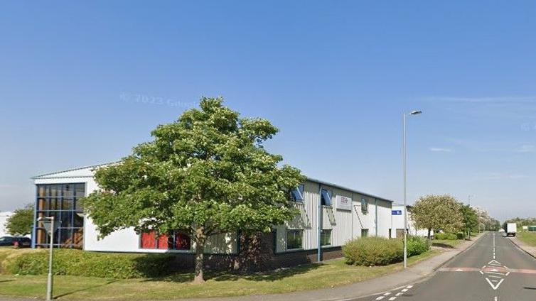 A Google Street View of the industrial estate. White, two storey-units are surrounded by grass, shrubs and trees. There is a large corner window on the left-hand side of the unit nearest the camera.