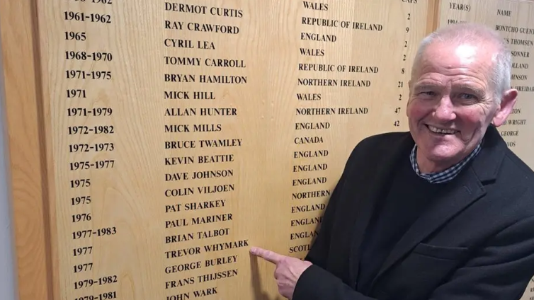 An elderly looking man with little hair and wearing a black coat over a jump. He is pointing at a wooden wall board which has names of Ipswich Town players written on it.