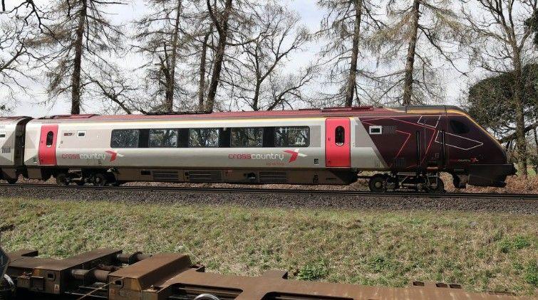 The cab and first carriage of a CrossCountry train travels on tracks in the countryside. In the background trees can be seen, with a grass bank visible in the foreground. 