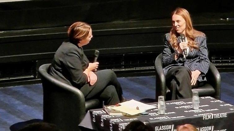 Kezia Dugdale and Jacinda Ardern are seated in armchairs on a stage during a discussion event, each holding a microphone. A table between them is covered with a black cloth featuring “Glasgow Film Theatre” branding