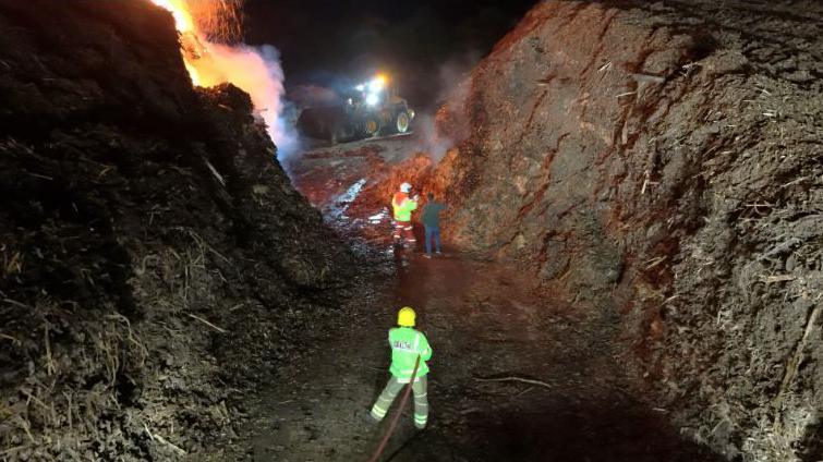 A firefighter with a hose standing between two high mounds of materials, with flames in the background. Another firefighter and a person in casual dress are standing closer to the fire.