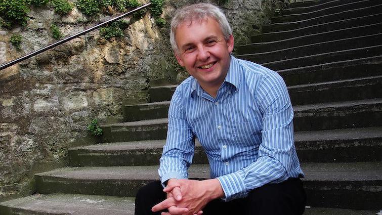 A smiling Colin Beveridge sitting on a long flight of stone steps. He is wearing a blue and white striped shirt and has his hands clasped.