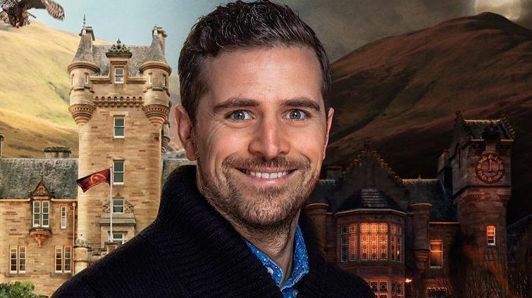 Alexander Dragonetti is smiling at the camera against a CGI background of a castle, half in daylight and half at night. He wears a black jumper, blue shirt and has brown hair and facial hair.