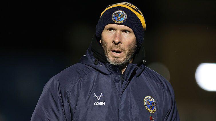 Shrewsbury Town manager Michael Appleton in his club puffer jacket and beanie walks along the touchline