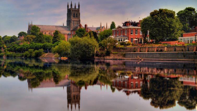 Worcester Cathedral on the banks of the river Severn, which is calm and reflecting the cathedral clearly. it is summer and there are trees and shrubs lining the river.