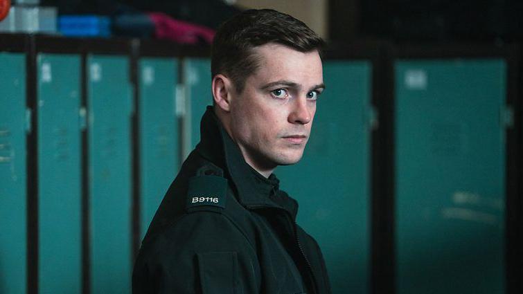 Nathan Braniff is pictured as his character Tommy Foster. He is stood with his right side facing the camera, with a neutral expression on his face. He is stood in front of teal coloured metal lockers and is wearing a green police uniform jacket.