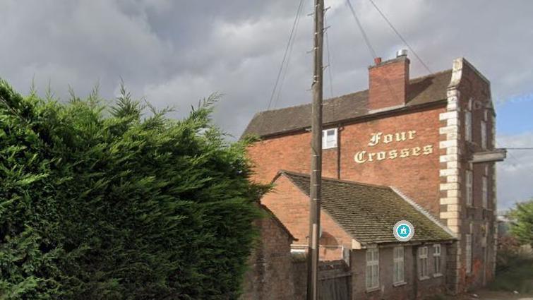 An old red brick building with a sign saying Four Crosses on the side. Nearests to the camera is a huge hedge, then a one storey building followed by a three story building with the sign.