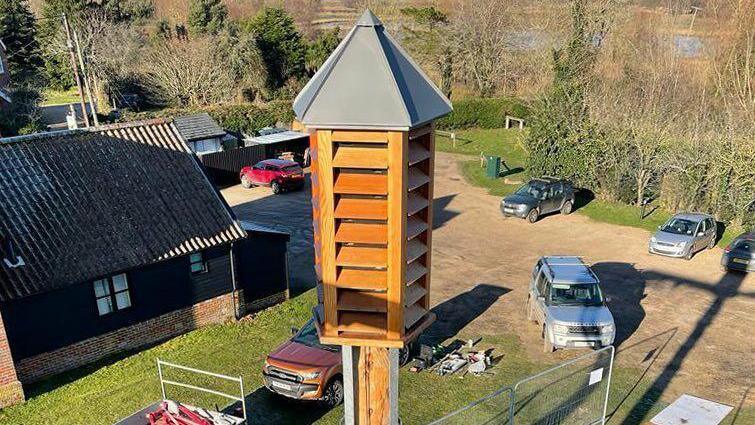 A high tower with wooden slats at the top is standing in a pub car park with vehicles and metal gates surrounding it