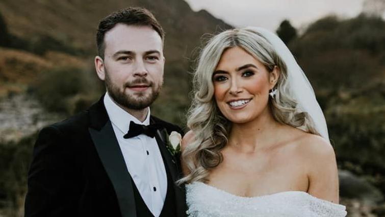 Paul and Siobhan McGowan pose for an outdoor photo on their wedding day.  Paul has short, dark hair and a beard and is wearing a black and white tuxedo with a black bow tie. Siobhan has long, curled blonde hair and is wearing a white, off-the-shoulder wedding dress and a white veil.