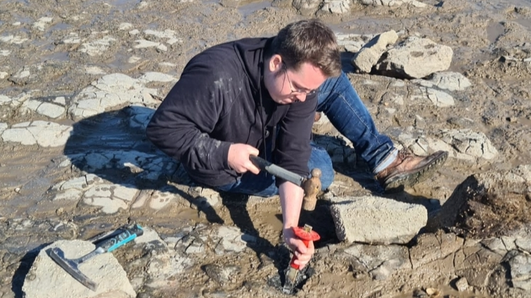 Owain Evans hammers a red handled chisel into the ground at a fossil field - he's wearing a black coat and blue jeans with splatters of mud on them