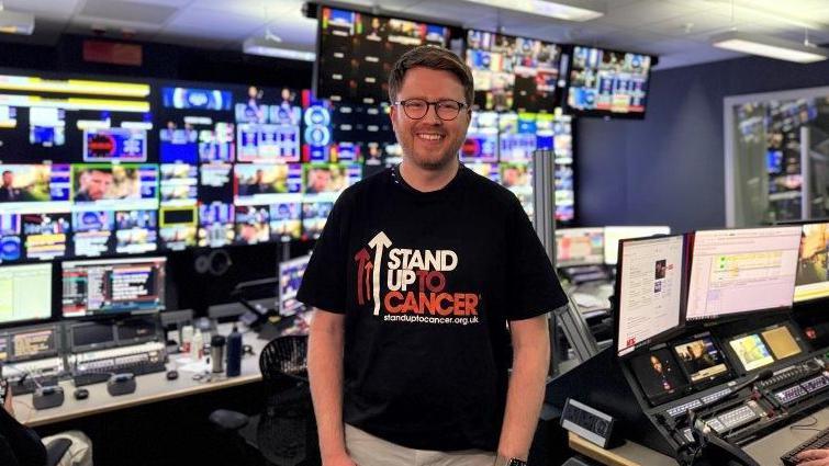 Jack wears a black Stand Up To Cancer shirt, and stands in the control room of a TV studio, backed by control desks and dozens of monitors. He has dark red hair and wears glasses.