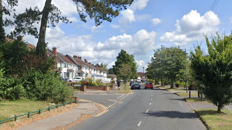 On a sunny day, the Google Streetview shot of Wagon Lane shows trees on both sides of the road, with a pedestrian ahead on a pavement and three cars can be seen in the distance