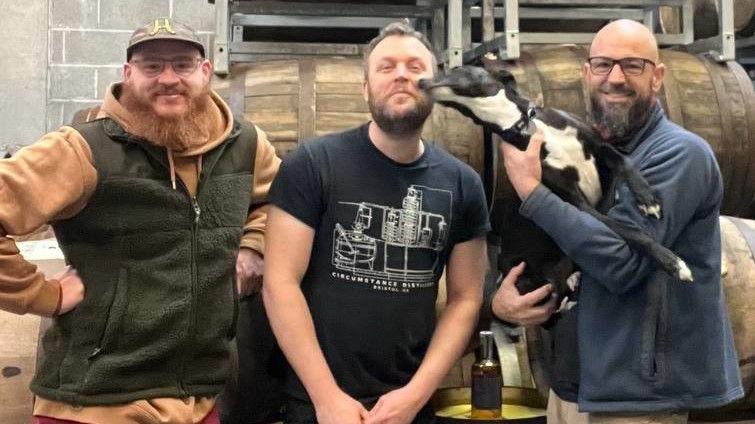 Three men with beards, one of them holding a whippet-style black dog with a white chest, pose in a line front of a set of wooden whisky barrels.