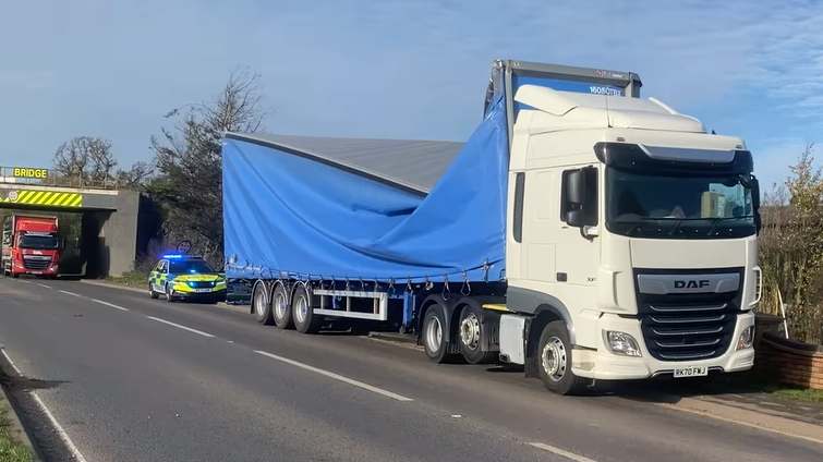 Damaged lorry with bridge visible in background and police car parked nearby