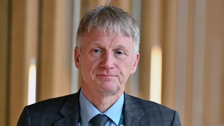 Ivan McKee walking in the Scottish Parliament building, wearing a grey checked suit, blue shirt and tie