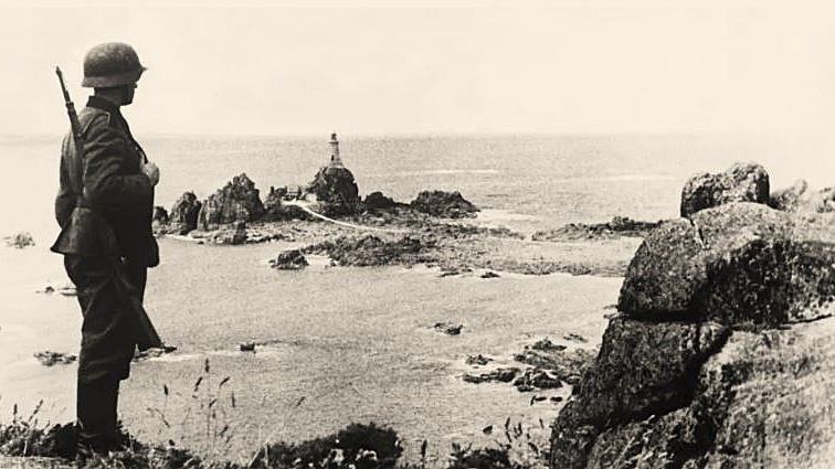 A German solider holding a rifle and wearing a tin helmet is standing on a cliff and looking out to sea