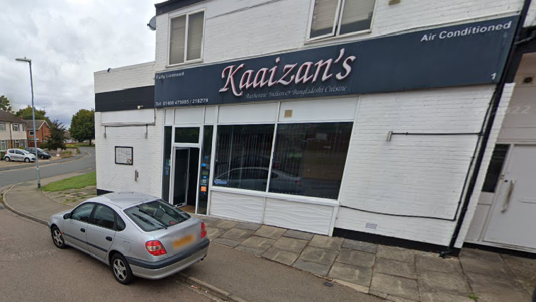 A white building with Kaaizan's written on a blue sign above a glass door. A grey car with its number plate blurred out is parked in front of the door.