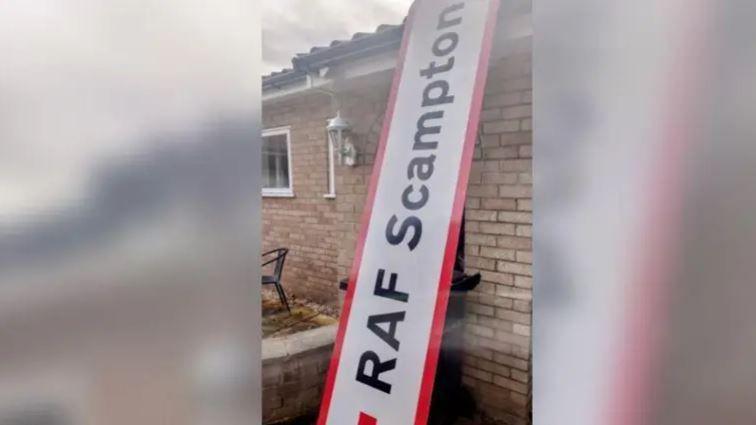 A road sign with ‘RAF Scampton’ written in black letters can be seen leant against a brick bungalow, it is oversized against the house. 