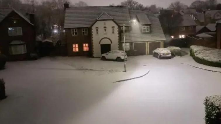 A snowy scene with a house and cars with a dusting of snow at night