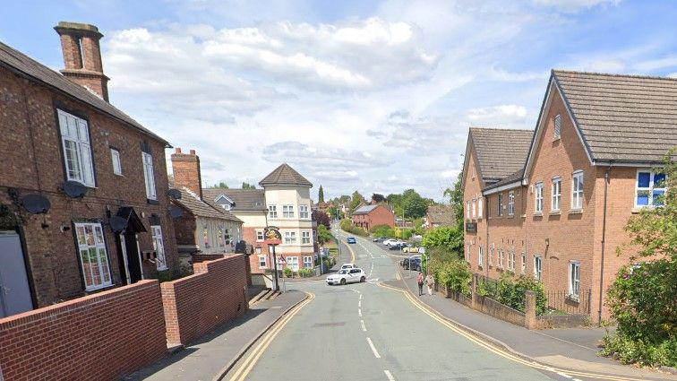 A road with houses and businesses including a pub at intervals along it. Halfway along, there is a mini-roundabout with a modern turreted building at the junction.