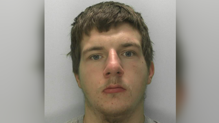 A mugshot of a 19-year-old with brown hair, staring blankly at the camera. He has spots on his chin. He has a grey sweatshirt on and is stood in front of a grey background.