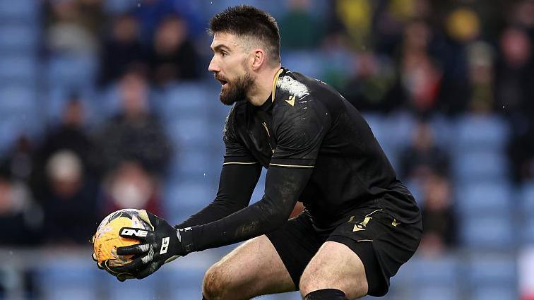 A bearded Matt Ingram wearing an all-black kit and black goalkeeping gloves with the ball in his hands during a game