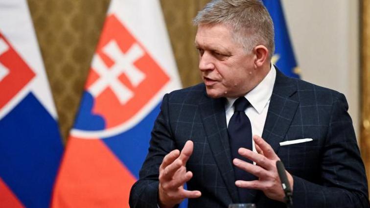 Robert Fico talks to reporters, wearing a dark suit and tie with a backdrop of two Slovak flags