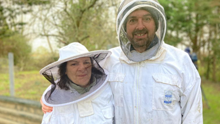 Luigi and Cherie Capozzoli smile at the camera, wearing white bee suits with the hoods up. Behind them a green field with trees can be seen.