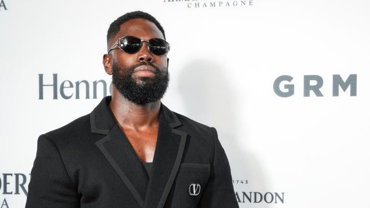 Ghetts at the GRM Gala. Ghetts wears a black suit over a black vest top, matched with dark sunglasses. He has a short beard and cropped dark hair. 